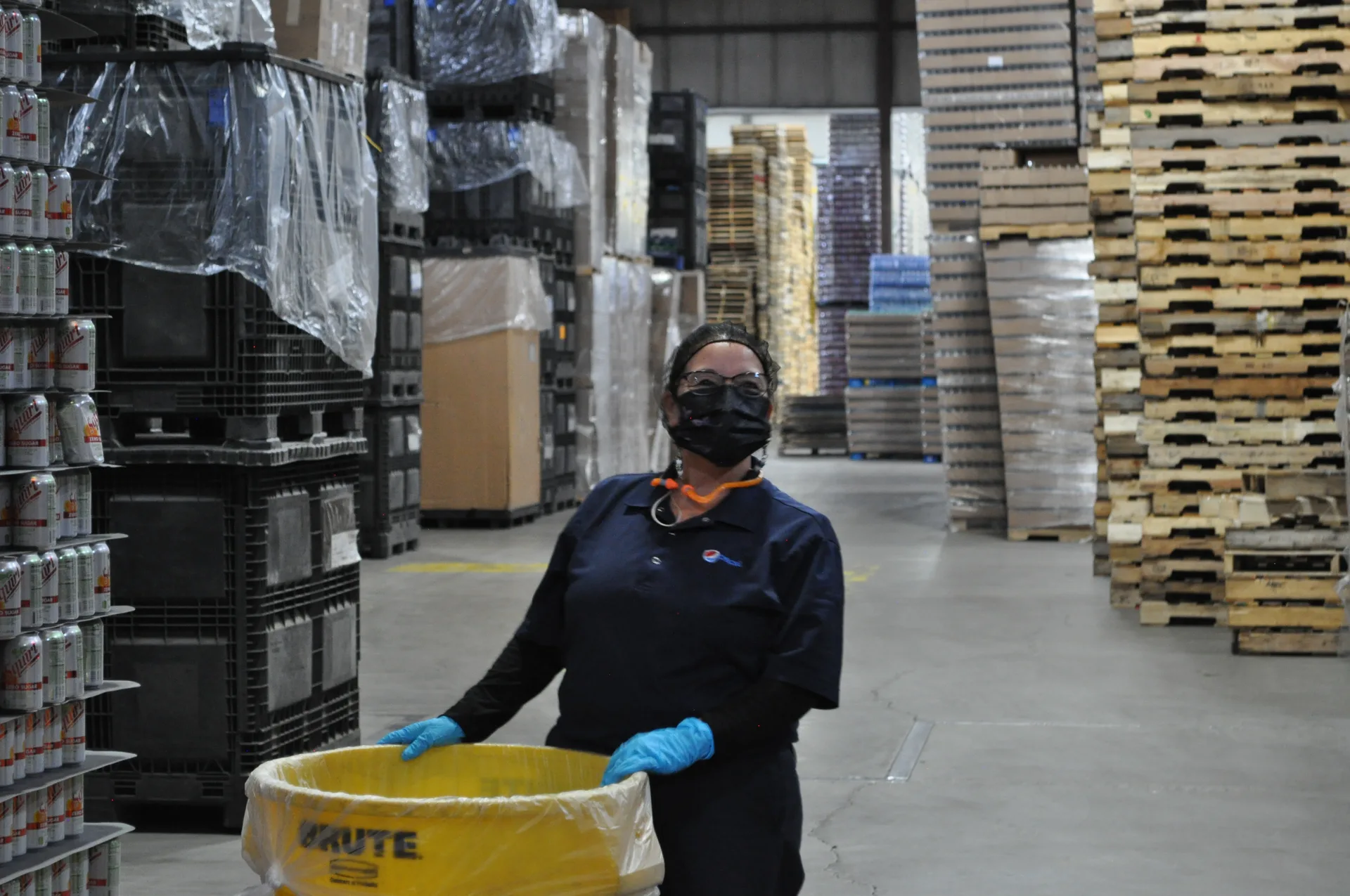 woman with a trash can and wearing a mask standing in a warehouse with empty pallets