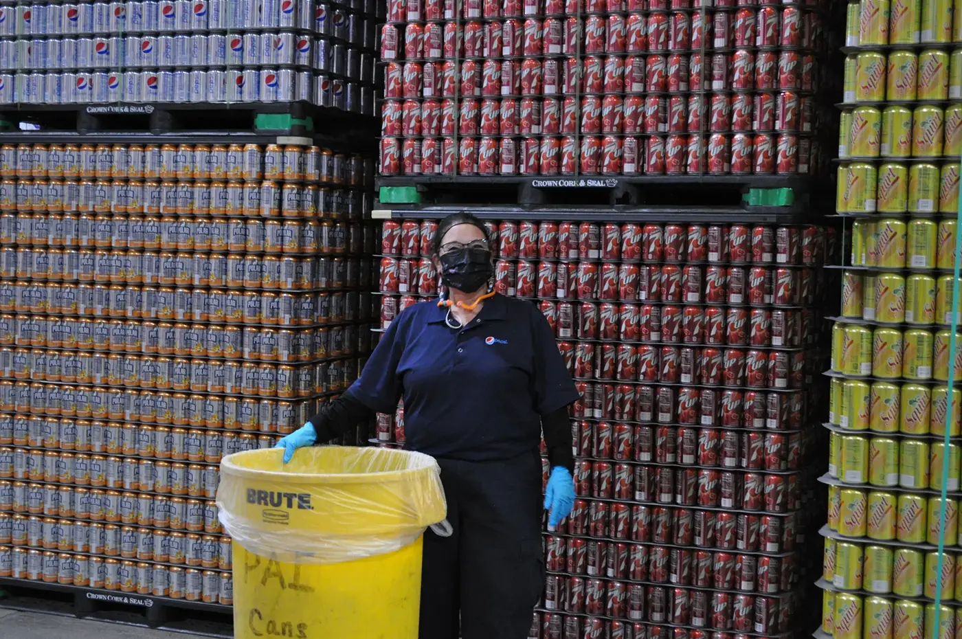 woman with a trash can and wearing a mask standing in front of pallets with cans of soda on them